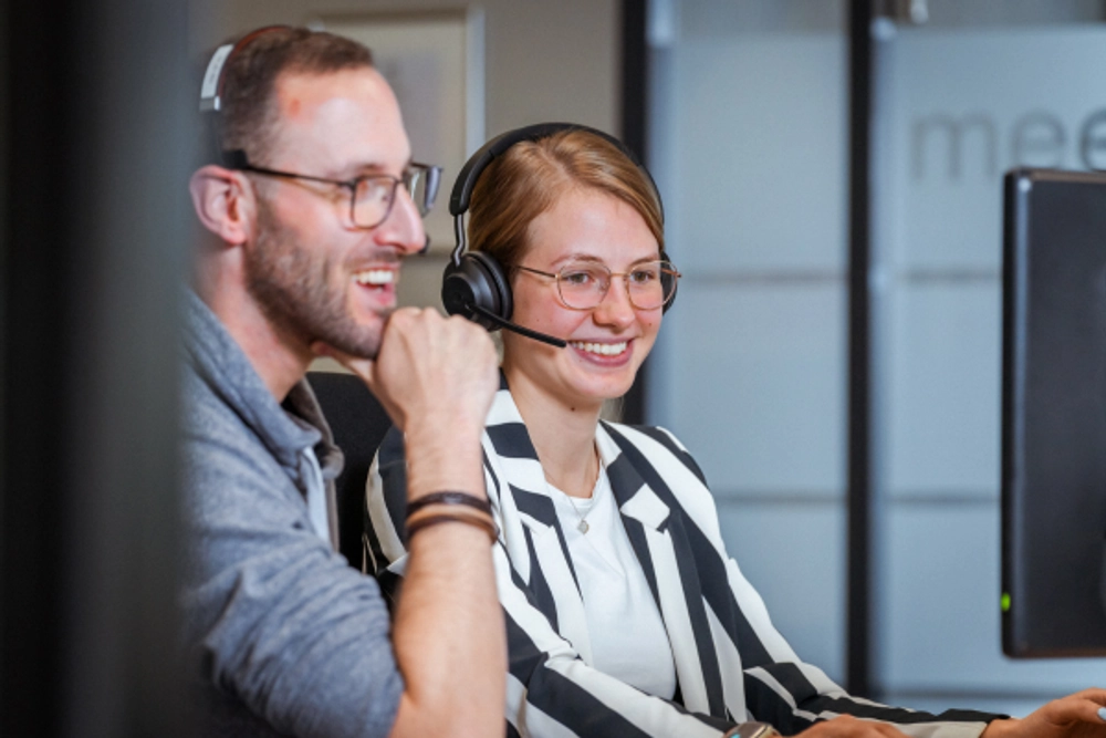 Das Bild zeigt zwei Personen, einen Mann und eine Frau, die beide Headsets tragen und vor einem Computer sitzen. Sie scheinen in einem Büro oder einem Callcenter zu arbeiten. Der Mann hat seine Hand am Kinn und scheint zuzuhören oder nachzudenken, während die Frau lächelt und in die Kamera blickt. Der Hintergrund ist unscharf.